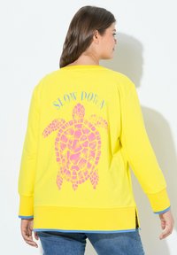 Sweatshirt jaune avec un graphique de tortue en rose au dos, et le texte "SLOW DOWN" au-dessus. Présente des bandes d'accent bleu aux poignets.