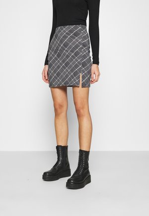Une femme portant un haut à manches longues noir, une mini jupe à carreaux gris avec une fente à l'avant, et des bottes plateformes noires à lacets, debout devant un fond uni.