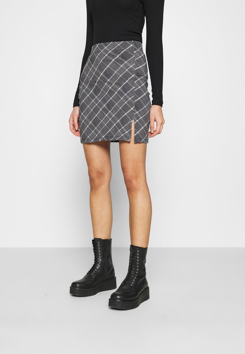 Une femme portant un haut à manches longues noir, une mini jupe à carreaux gris avec une fente à l'avant, et des bottes plateformes noires à lacets, debout devant un fond uni.
