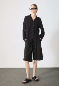 Svart långärmad skjorta med knappar, kombinerad med svarta knälånga shorts och svarta slidesandaler, vilket skapar en monokrom look.