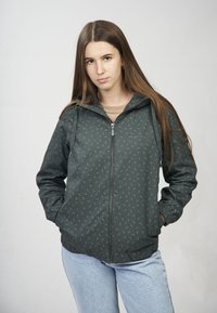 Jeune femme aux longs cheveux bruns portant une veste zippée à motifs vert foncé et un jean bleu clair, debout les mains dans les poches.