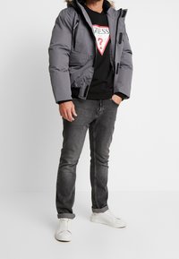 Giacca a guscio grigia con cappuccio rimovibile, tasche con zip, felpa nera, jeans grigi e sneakers bianche. Presenta cuciture a contrasto e zip.