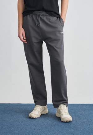 Persona che indossa pantaloni grigi scuri con coulisse e scarpe da ginnastica beige chiaro, in piedi su un tappeto blu contro una parete bianca.