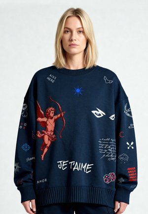 Femme blonde portant un sweat-shirt bleu marine surdimensionné avec un cupidon, des yeux, des dés et des textes liés à l'amour comme "Je t'aime" et "Amour" dans différentes polices.