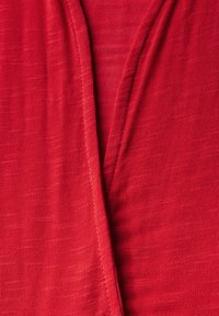 Röd tyg med en slät textur, som har en djup V-formad halsringning och overlappande lager. Materialet verkar lätt och något genomskinligt.