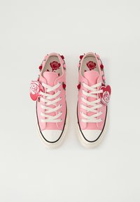 Zapatillas de lona rosas con adornos florales y etiquetas en forma de corazón, con cordones blancos y suelas de goma negra.