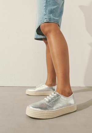 Personne portant des baskets à plateforme métalliques argentées et un short en denim clair arrivant au genou, debout sur un sol lisse devant un mur blanc.