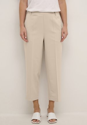 Beżowe, przycięte, proste spodnie noszone z białymi sandałami mule z odkrytymi palcami oraz białym bezrękawnikiem.