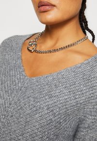 Grauer gestrickter Pullover mit V-Ausschnitt, kombiniert mit einer silbernen Kettenhalskette mit herzförmigen Hardware-Details.