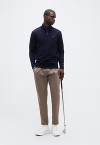 Homme debout les mains dans les poches, tenant un club de golf, portant un pull bleu marine, un pantalon beige et des baskets blanches sur fond uni.