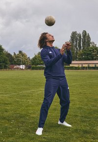 Chándal atlético de la marina con acentos blancos, jugador en movimiento, equilibrando un balón de fútbol sobre su cabeza en un campo de hierba.