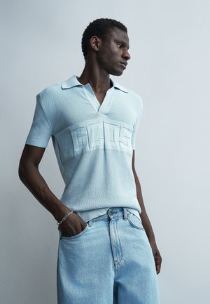 Άνδρας που φοράει ανοιχτό μπλε πλεκτό πόλο με ανάγλυφο λογότυπο και ανοιχτό μπλε τζιν, στέκεται με το ένα χέρι στην τσέπη, κοιτάζοντας πλάγια σε απλό φόντο.