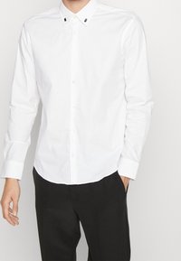 Chemise blanche à manches longues en coton, avec un col boutonné, de petits accents noirs sur le col et un ourlet arrondi.