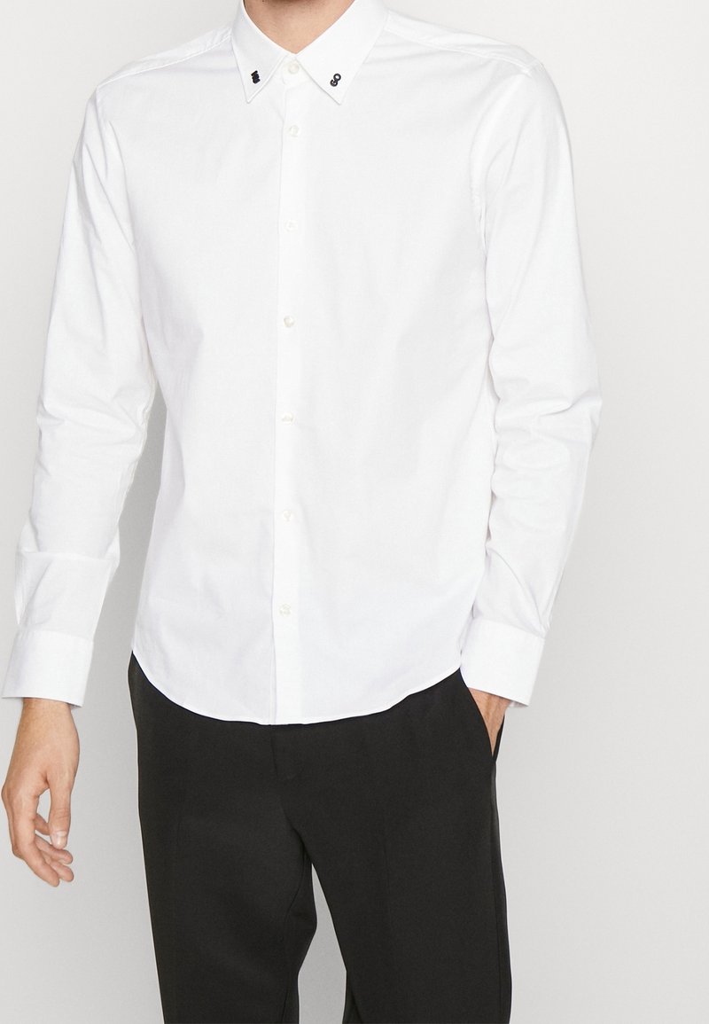 Chemise blanche à manches longues en coton, avec un col boutonné, de petits accents noirs sur le col et un ourlet arrondi.