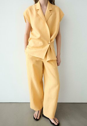 Kvinde iført løs, gul ærmeløs blazer og bukser med vide ben kombineret med sorte thongsandaler, stående mod en ensfarvet hvid væg.
