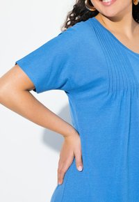 Niebieska bluzka z krótkim rękawem i okrągłym dekoltem, z pionowymi zakładkami wzdłuż klatki piersiowej, wykonana z gładkiego, lekkiego materiału.