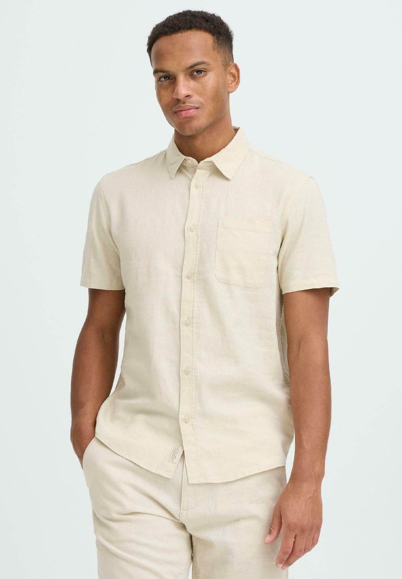 Chemise beige en lin à manches courtes avec col boutonné, poche poitrine simple et coupe décontractée. Le tissu présente une texture douce et texturée.