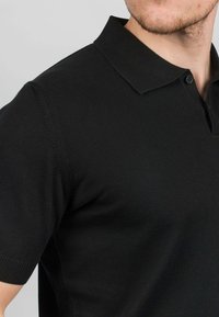 Svart polo-tröja med ribbad textur, korta ärmar och klassisk krage. Har en knappslå på framsidan.