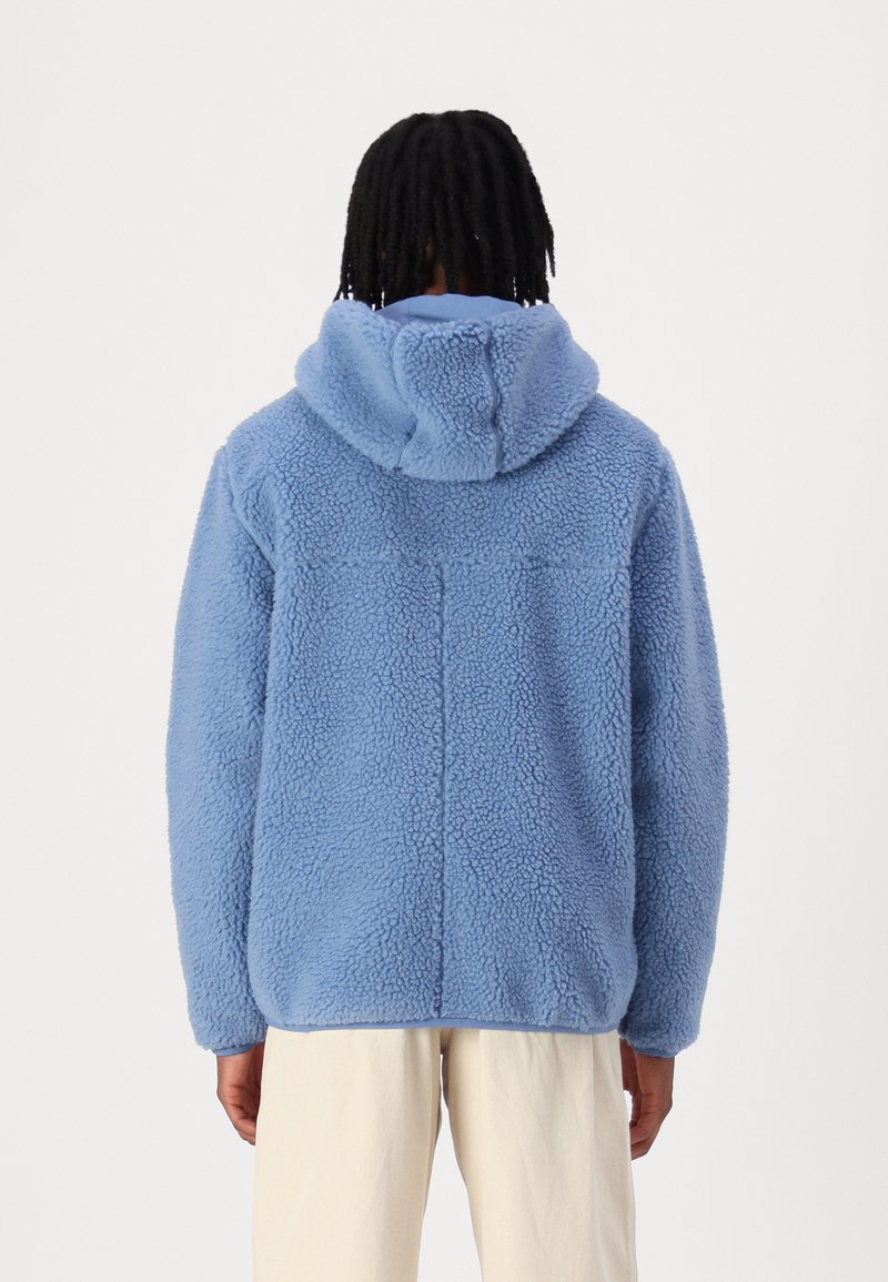 Blaue Fleecejacke mit Kapuze, die eine strukturierte, flauschige Oberfläche und einen lässigen Schnitt aufweist. Die Rückseite zeigt ein einfaches Design mit Nahtdetails.