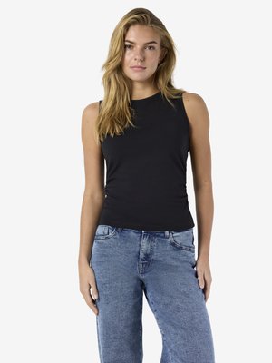 Joven mujer con cabello rubio largo, vestida con una blusa negra sin mangas y jeans azules, de pie frente a un fondo blanco liso.