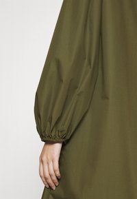 Main reposant à côté du bras d'un vêtement vert olive avec un poignet élastique froncé sur fond blanc uni.