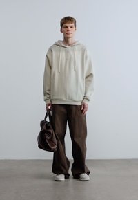 Μπεζ φούτερ με κάθετη ρίγα, κορδόνια και λογότυπο. Καφέ παντελόνια με φαρδιά ποδάρια. Το μοντέλο κρατά καφέ δερμάτινη τσάντα, φοράει λευκά αθλητικά παπούτσια.