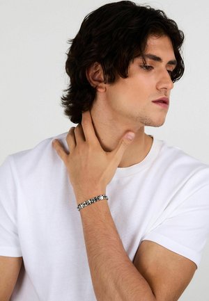 Giovane uomo con capelli scuri e mossi, in t-shirt bianca, che si tocca il collo con la mano sinistra, indossando un braccialetto a scacchi nero e argento.