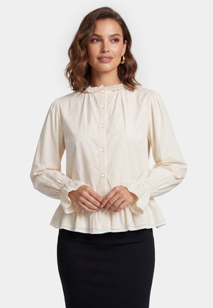 Vrouw draagt een crèmekleurige blouse met lange mouwen en knopen, met ruches aan de manchetten en kraag, gecombineerd met een zwarte rok, staand met handen samengevouwen.