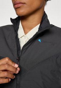 Giacca nera con colletto alto, chiusura a zip, pannello testurizzato e logo blu. Il modello indossa una camicia color crema sotto.