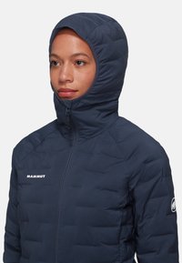 Marinblå isolerad jacka med huva, kviltad design, framdragkedja och "MAMMUT"-logotyp p�å bröstet. Mjuk textur, kompakt form.