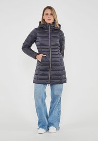 Donna in piedi con un piumino nero con cappuccio e zip, jeans a campana blu e sneakers bianche su uno sfondo bianco uniforme.