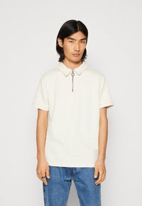 Short-sleeve polo shirt in cream with a textured pattern, featuring a zip-up collar and ring pull, paired with blue denim jeans.