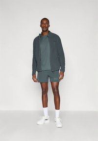 Muški sportski komplet: tamnozelena jakna na patentni zatvarač, odgovarajuća majica kratkih rukava i kratke hlače, upareni s bijelim sportskim cipelama i čarapama do teleta.