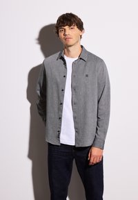 Camicia button-up grigia con una sottile texture, indossata sopra una maglietta bianca. Il modello si trova di fronte a uno sfondo semplice, indossando jeans scuri.