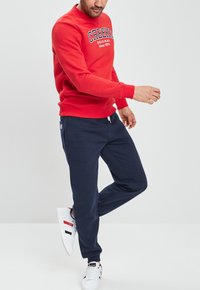 Sudadera roja con el texto "CREEKS ORIGINAL Desde 1974", combinada con pantalones de chándal azul marino y zapatillas blancas con rayas rojas y negras.