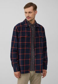 Marineblauwe flanellen jas met rood geruit patroon, twee borstzakken en knoopsluiting. Gedragen over een geruit shirt en gecombineerd met lichte broek.
