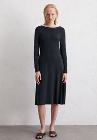 Schwarzes langärmliges Kleid aus weich material, mit einem Bootsausschnitt und einer fließenden A-Linien-Silhouette. Getragen mit schwarzen Slipper-Sandalen.