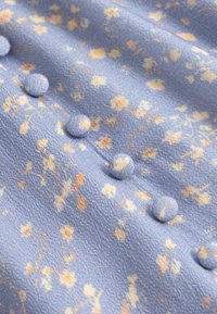 Tissu floral violet clair avec de petites fleurs jaunes et beiges, présentant une rangée de boutons ronds recouverts le long d'une couture.