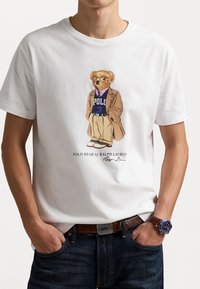 Fehér pamut póló, amelyen egy szemüveget viselő mackó illusztráció látható, öltönykabáttal és "Polo" pulóverrel, bézs nadrággal és sportcipővel.