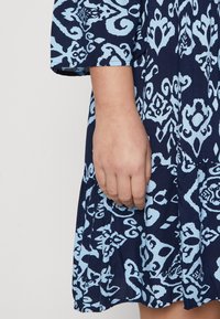 Mörkblå klänning med ljusblå abstrakt mönster, korta ärmar, plisserad kjol och flödande tyg. Närbild av en hand som vilar på klänningen.