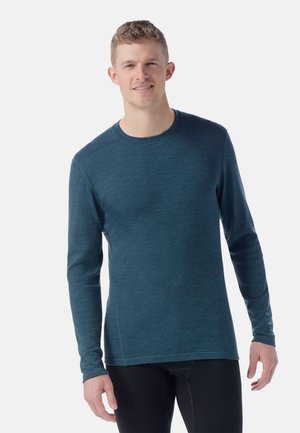 Langærmet skjorte i teal stof med rund halsudskæring, glat tekstur og subtile syninger på ærmerne og kanten.