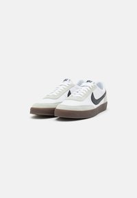 Zapatillas Nike con parte superior de cuero blanco y acentos de ante gris, logo de swoosh negro, suela de goma marrón, diseño con cordones y punta redondeada.