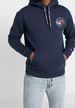 Mann trägt dunkelblauen Hoodie mit Fronttasche und Logopatch, kombiniert mit hellblauen Jeans, Hände in den Taschen.