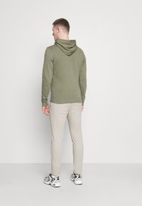 Olive green hoodie with a kangaroo pocket and a drawstring hood, paired with light beige joggers and sneakers, viewed from the back.