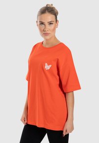 Oranžové bavlněné tričko s krátkým rukávem, na levé straně hrudi s malou bílou grafikou motýla. Uvolněný střih, hladká textura.