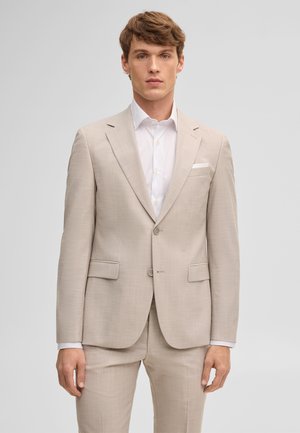 Uomo con abito beige a due bottoni, camicia bianca e fazzoletto bianco nel taschino, in piedi davanti a uno sfondo grigio chiaro uniforme.