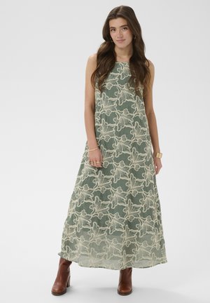 Jeune femme portant une robe maxi verte sans manches avec une broderie florale blanche et des bottines marron à talons, debout devant un fond uni.
