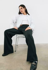Wit en zwart gestreepte oversized shirt, witte crop top, zwarte wijde broek, zwarte loafers met gouden hardware, zittend op een draadstoel.