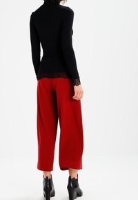 Kvinde iført en sort langærmet top med blonde detaljer, røde bred-bens beskårne bukser og sorte hælede ankelstøvler, stående med ryggen til.