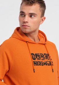 Felpa arancione con testo nero "OBERE MENSWEAR" sul davanti. Presenta un cappuccio con cordoncino e un tessuto textured.